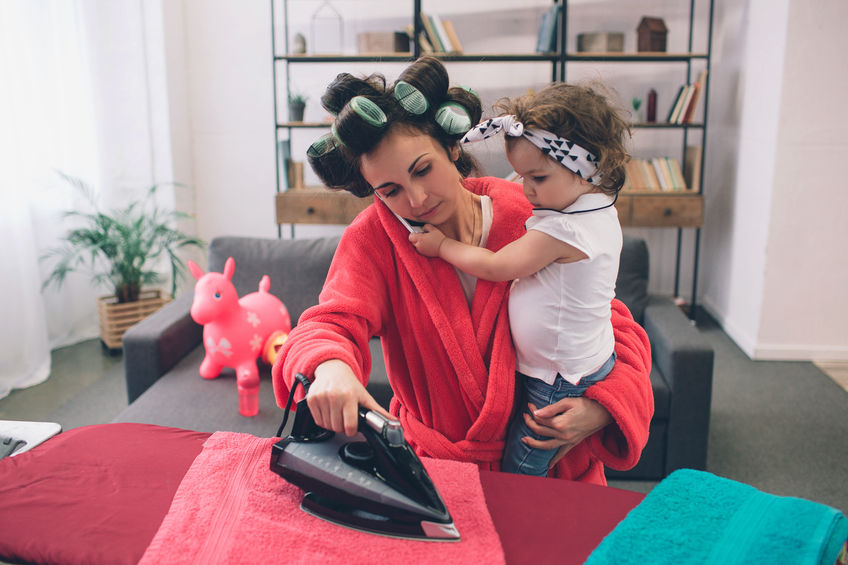mother and baby together engaged in housework Ironing clothes . Housewife and kid doing homework. Woman with little child in the living room. Homemaker doing many tasks while looks after her infant.