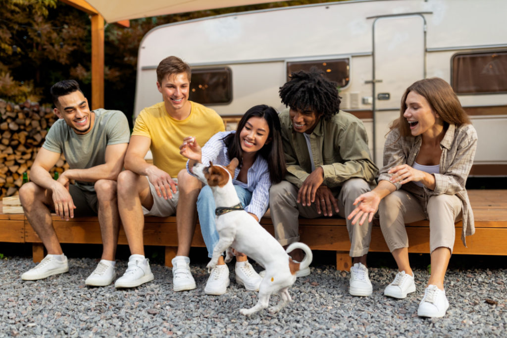 Happy multiethnic young friends sitting near RV, playing with pet dog, enjoying autumn vacation on campsite
