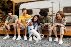 Happy multiethnic young friends sitting near RV, playing with pet dog, enjoying autumn vacation on campsite